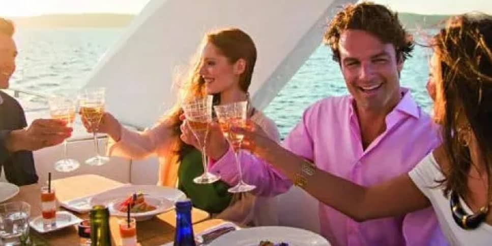 People on yacht enjoying drinks: A photo of people on a yacht, with glasses and bottles visible on a table in the foreground, and the ocean and horizon visible in the background.
