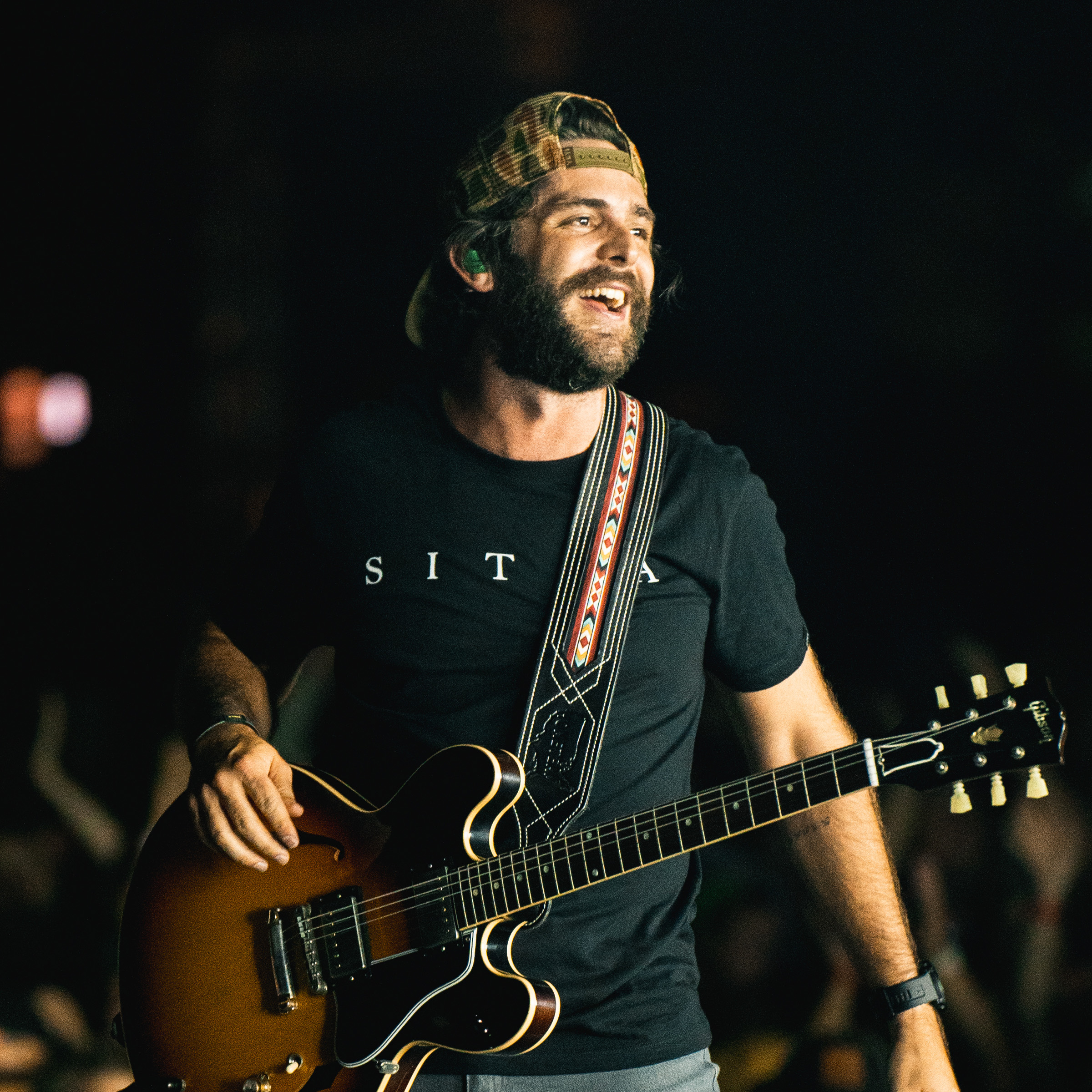 Country music artist Thomas Rhett performed on stage, holding a microphone and singing into it. He wears a denim jacket and a cowboy hat, while the crowd cheers in the background."